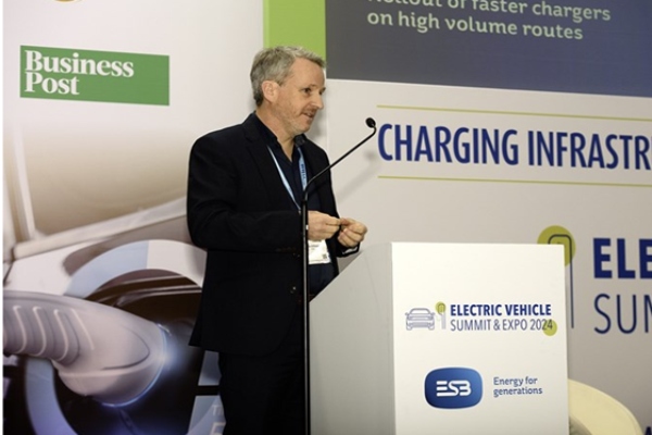 white man with grey hair, wearing a dark suit standing at podium with branding exhibition stand behind them