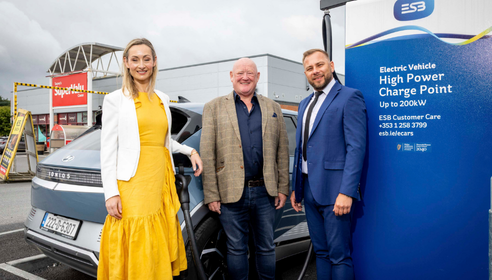Three people standing in front of a grey car and beside a large EV charging infrastructure.