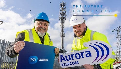 Two men wearing yellow high vis jackets and hard hats standing at a telecommunications site with a Tower in the background and both holding branded signs.