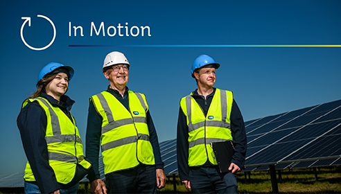 Photograph of a woman and two men in high-vis jackets and hard hats standing in front of large solar panels in a field, with text overlay reading 'In Motion'