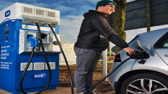 A man with a black jacket, black hat and grey jeans holding an electric vehicle tethered cable with a charge point in the bacground.