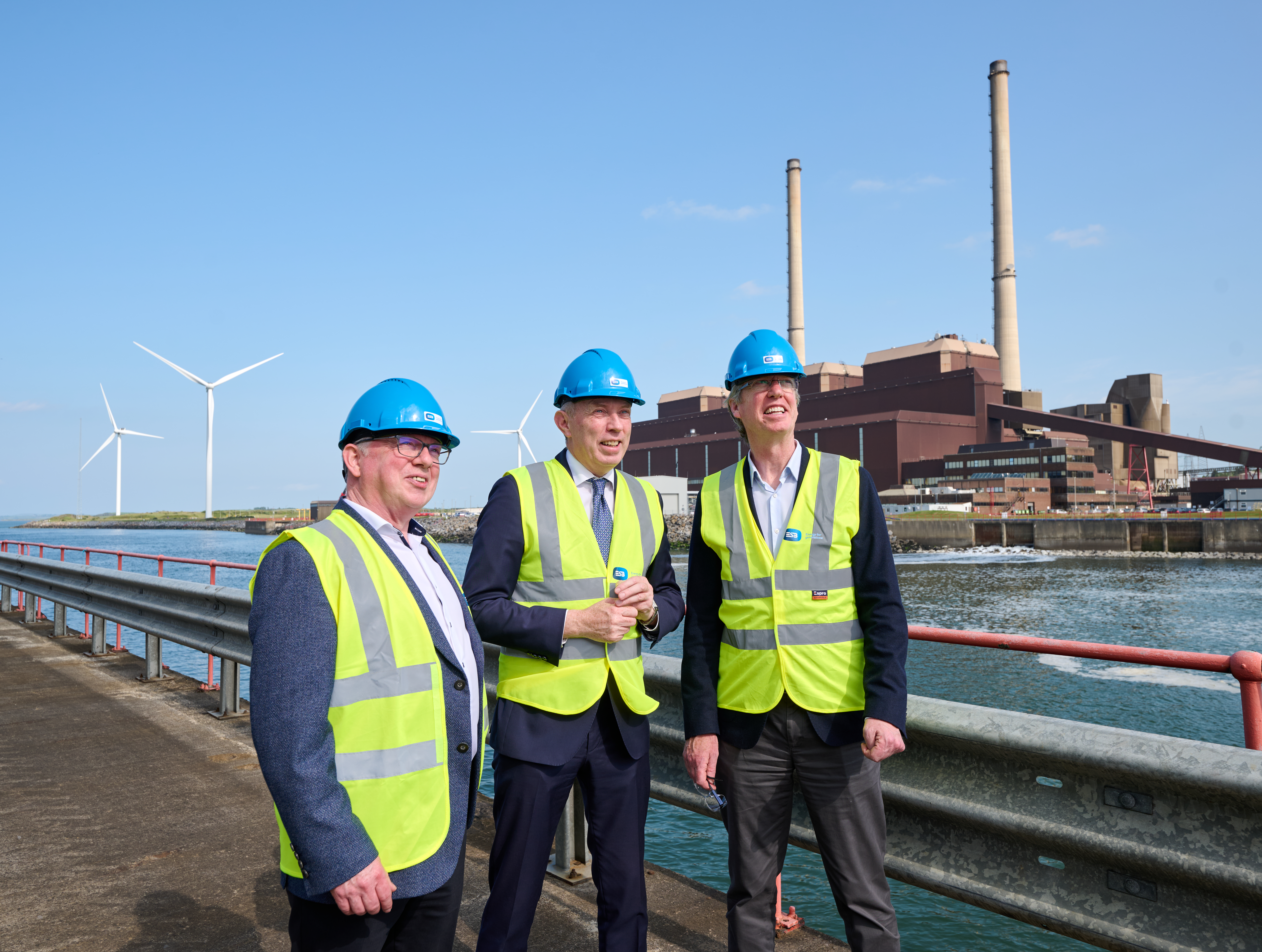 Three men in business attire wearing ESB-branded high-visibility vests and hard hats stand in front of Moneypoint power station and wind turbines