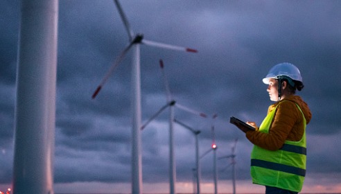 Photograph of woman in hard hat and high-vis vest looking at a tablet in front of a row of wind turbines