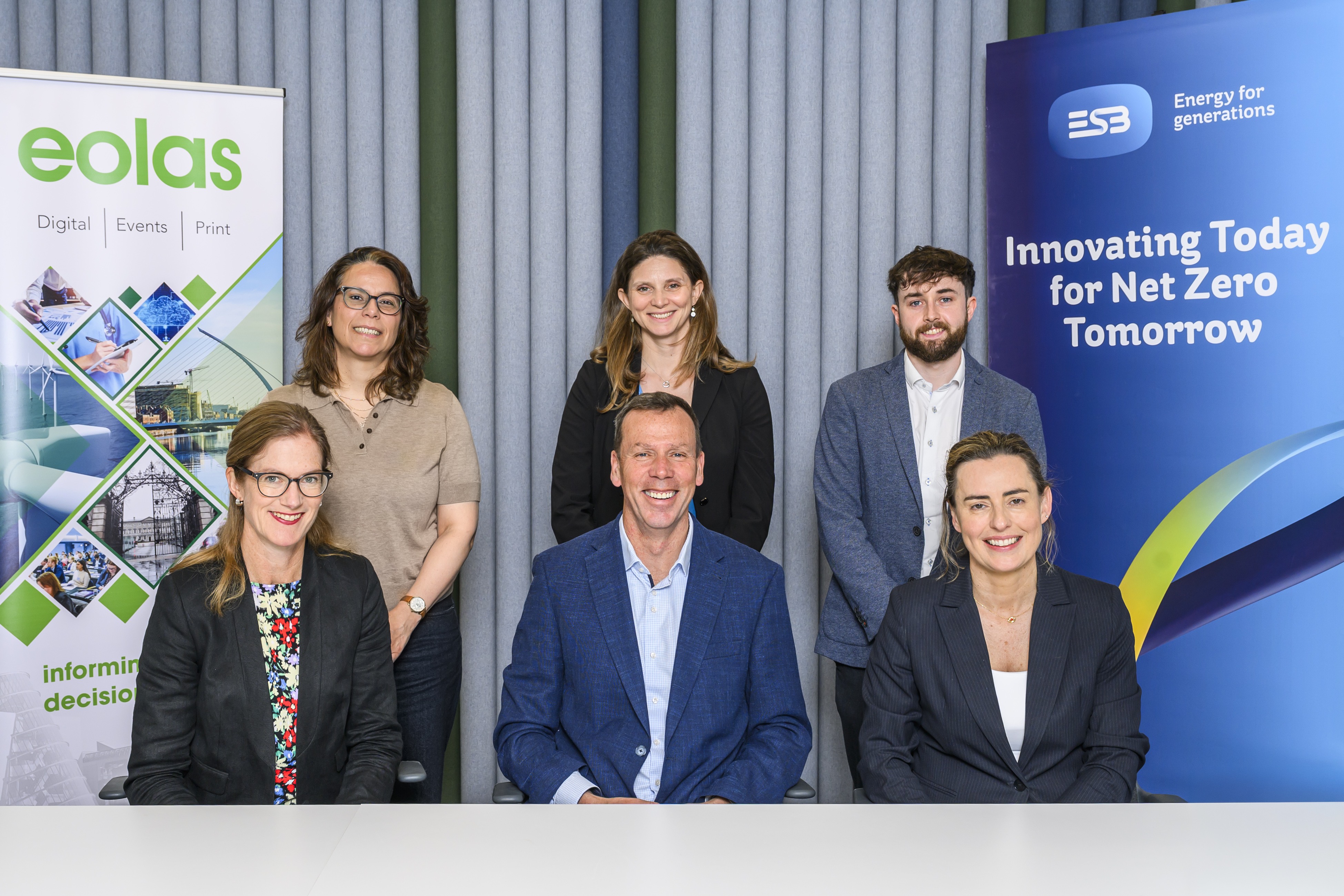 Group of six people in business attire posing for photograph, with two promotional banners in the background for Eolas magazine and ESB 'innovation for net zero'.
