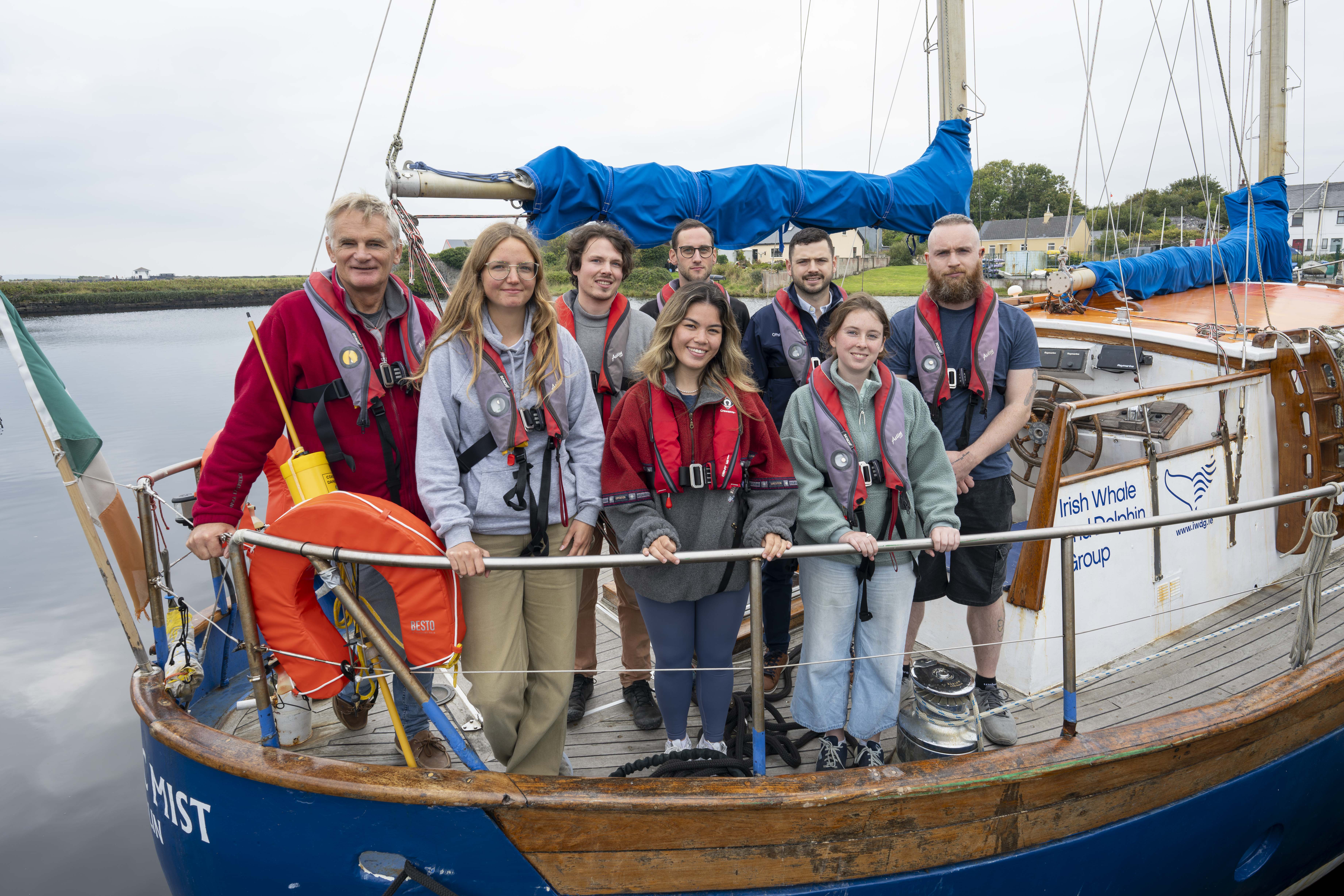 A group of five men and three women are standing facing the camera on a white and blue boat. They are all wearing red life jackets. The boat is surrounded by calm waters, and a grey sky.