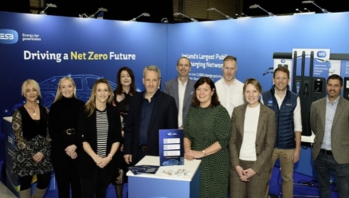 Group of 11 people posing in front of a blue backdrop. Small stand in center foreground with blue display sign.