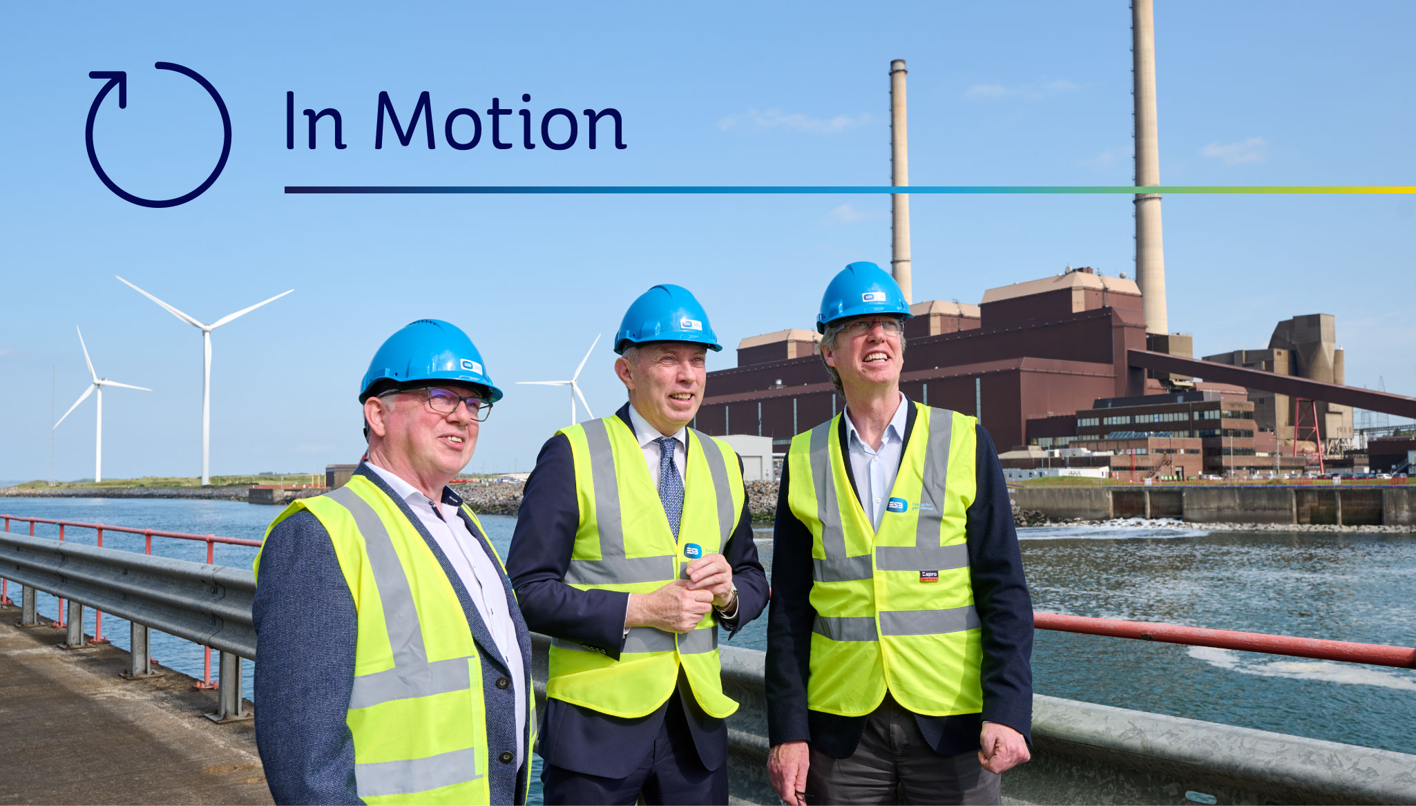 Photograph of three men in business attire and ESB-branded safety vests and hard hats, with power plant and wind turbines in the background. The words 'In Motion' appear at the top of the picture.