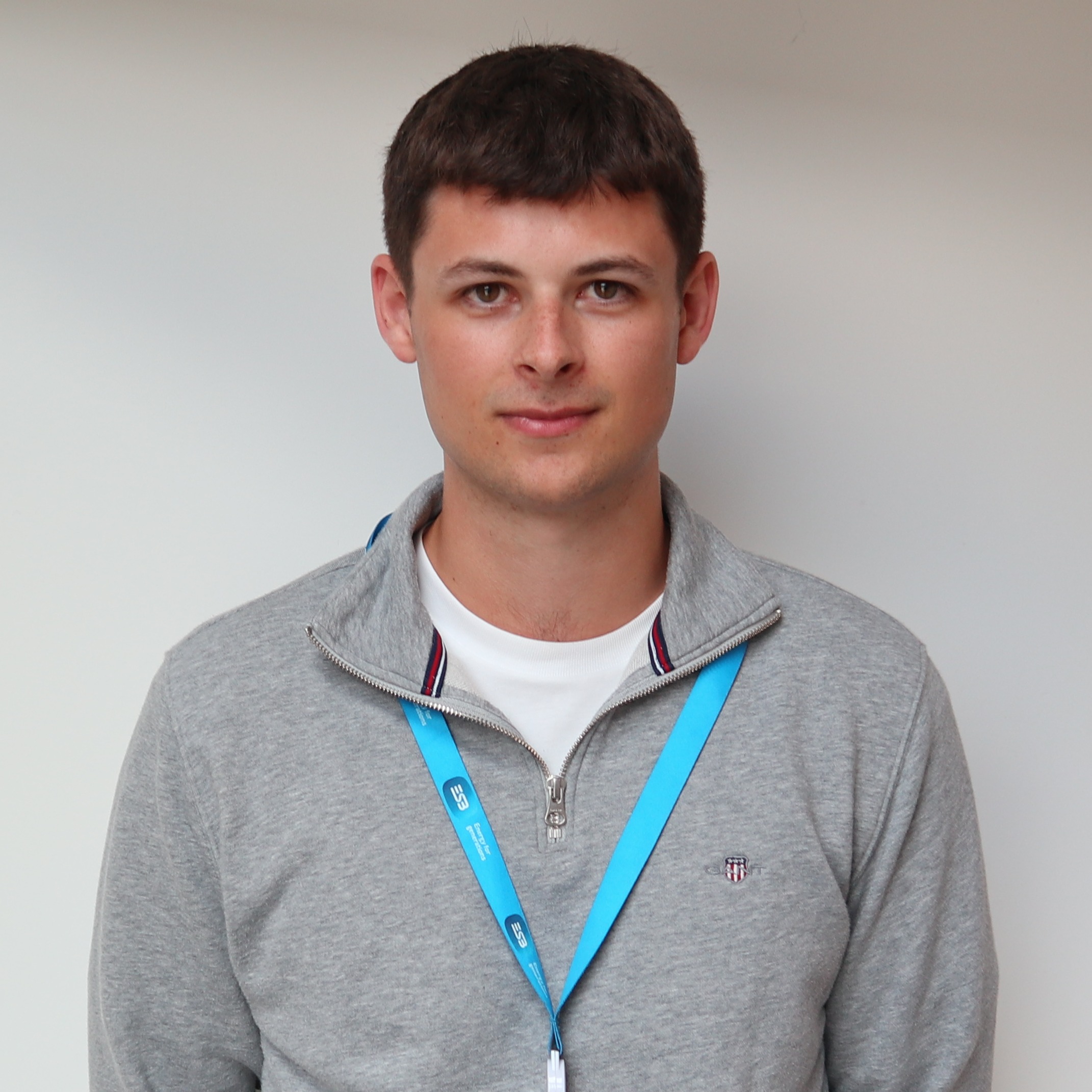 A person stands against a white wall wearing a gray quarter‑zip sweater, beige pants, and a blue lanyard with an ID badge