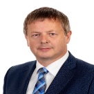 White man with grey and black hair wearing a black suit jacket, white shirt and striped blue tie.