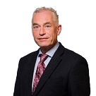 White man with grey hair and wearing a black suit jacket, white shirt and colourful tie.