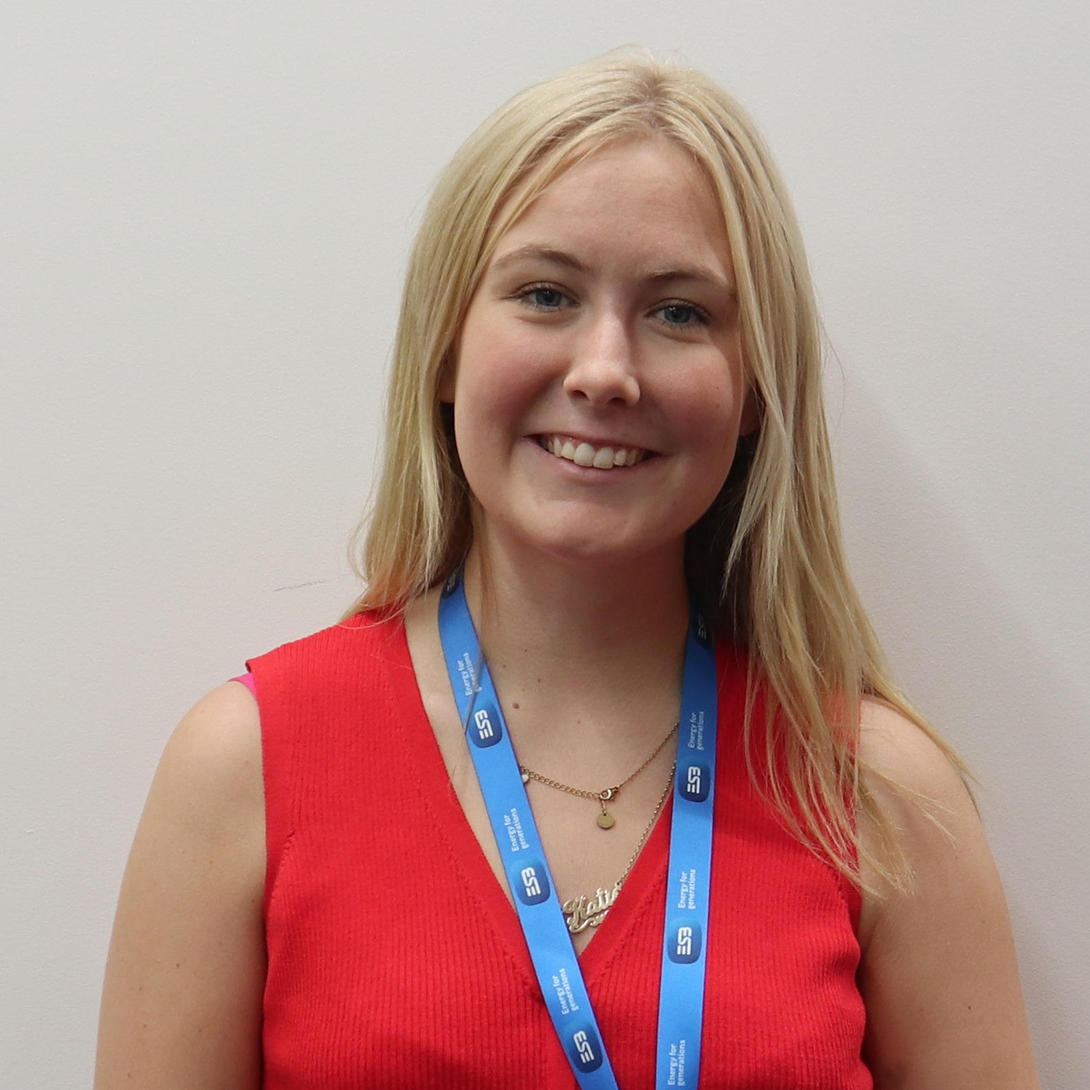 A person stands against a white wall wearing a red top, black pants, and a blue lanyard with an ID badge.