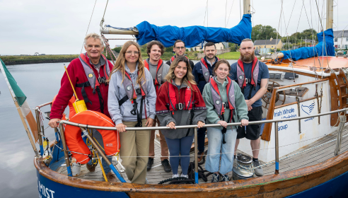 A group of five men and three women are standing facing the camera on a white and blue boat. They are all wearing red life jackets. The boat is surrounded by calm waters, and a grey sky.