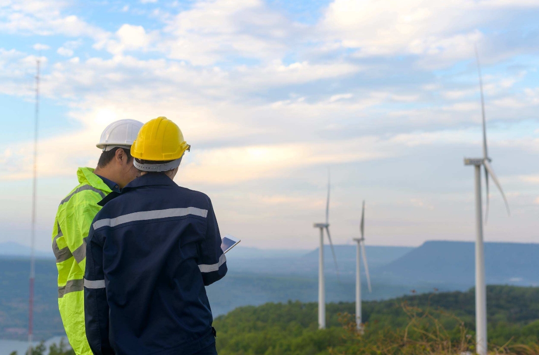 Two workers inspecting a wind farm while viewing information on a tablet, with wind turbines visible in the distance.