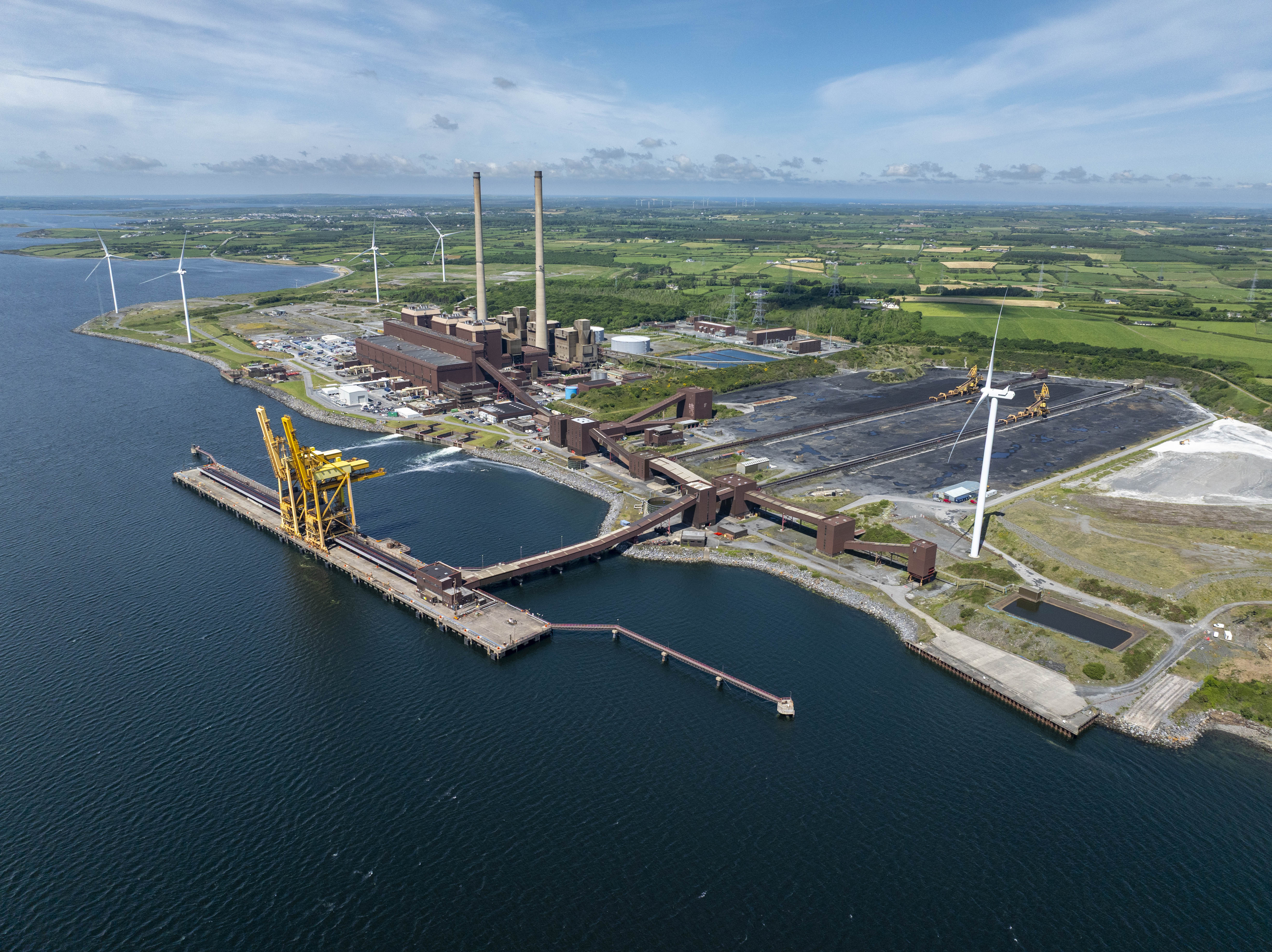 Aerial shot of power station on the coast, featuring large smokestack chimneys and coal yard as well as multiple wind turbines