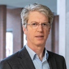 White man with grey hair, wearing glasses and a blue shirt with black jacket