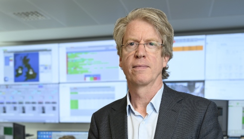 Man with short hair, wearing glasses and a blue shirt and dark navy jacket in front of large coloured screens