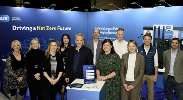 Group of 11 people posing in front of a blue backdrop. Small stand in centre foreground with blue display sign.