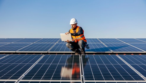 Man wearing a white hard hat and orange high vis jacket with an ipad in hands and crunching down on solar farm panels