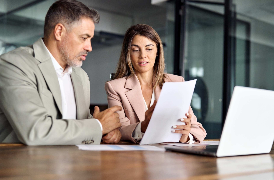 Two colleagues discussing a document at a meeting table with a laptop in an office.