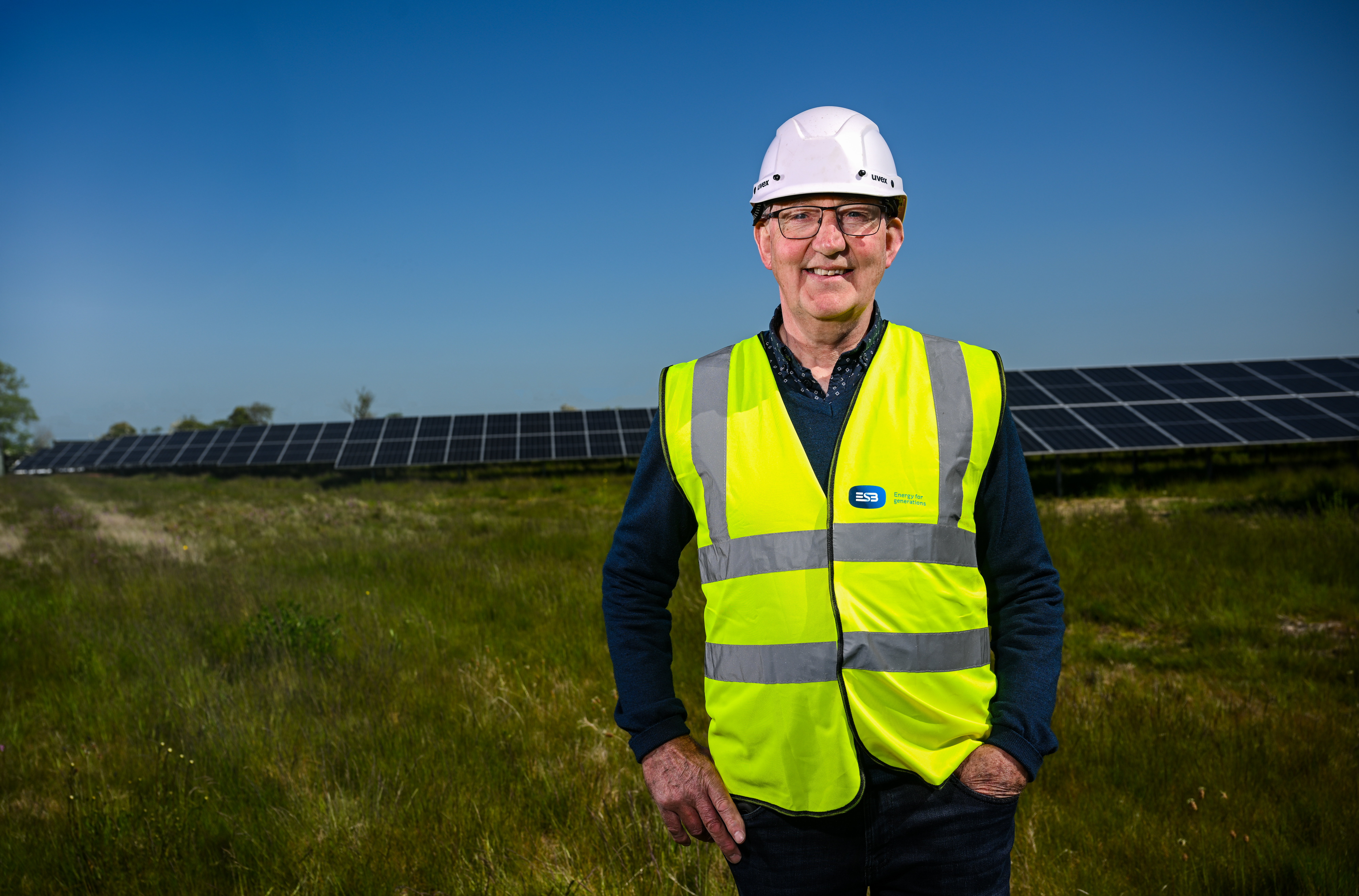 Photograph of a man in a high-vis vest and helmet standing in front of solar farm in a field