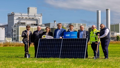 A group gathers on a grassy field outside an industrial site, posing with shovels and solar panels with branded signs.
