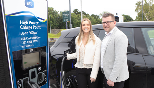Woman and man standing at black car with her hand charging the EV and large charging infrastructure to the forefront of the picture
