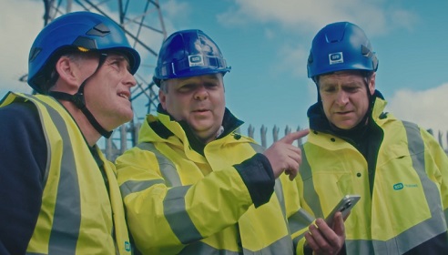 Three men wearing high vis and protective clothing, one has a phone in a hand and they are at a telecommunications site.