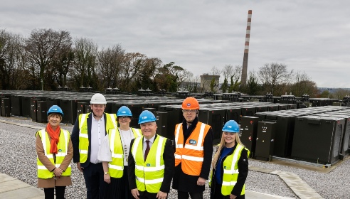 Six people wearing coloured hard hats and high visibility jackets standing in front of a battery energy storage plant