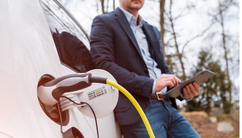 Man standing holding an ipad leaning against a car while it is charging