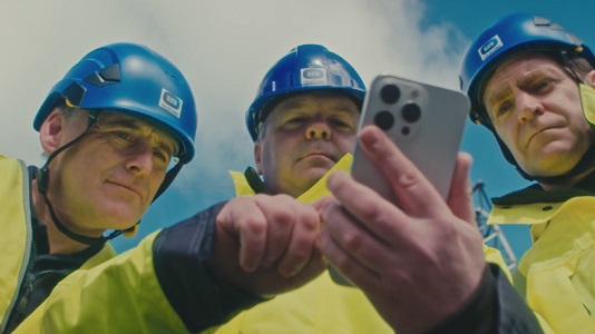Close up image of 3 ESB Telecoms men wearing high vis and protective hats, looking closely at a phone.