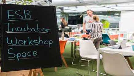 An office with two people in background with table and chairs with a sign that says 'ESB Incubator workshop Space.'