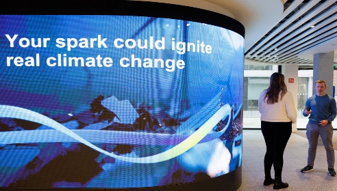 A screen at head office with the words 'Your Spark could ignite real climate change' and there are two people standing by the screen