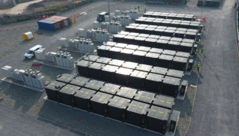 A row of seven battery electricity generational storage facilities on a large grey site with a white van and blue shed in the background.