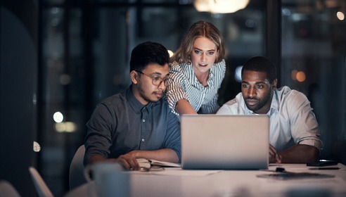 Two men and one woman working and looking a laptop. Two men are sitting and the woman is standing up and pointing at the laptop.