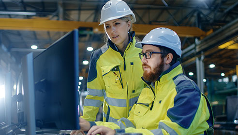A man and a woman wearing high vis and protective clothing in a technical work setting, both are looking at a laptop.