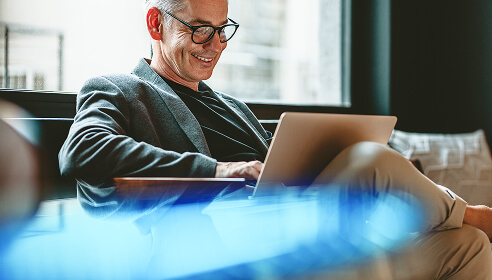 A man sitting down with grey hair and glasses with a laptop in front of him and a blue wave style decorative image crossing the picture