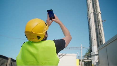 Man with his back to the photo, taking a photo of a telecommunications tower. He is wearing a high vis and a hard hat, and the sky is very blue.