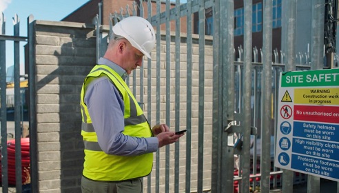 Image of a man at a gate which is the entrance to a telecommunications site. He is in high vis and wearing protective gear, there is a safety sign on a fence and the man is looking at this phone.