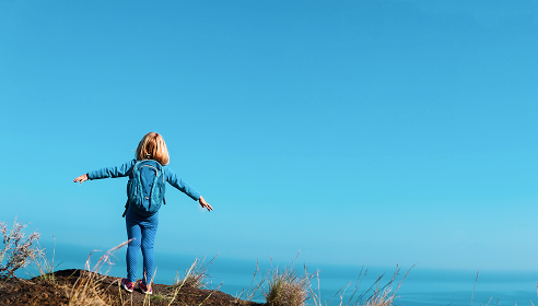 A girl standing on a hill with hands outstretched and looking out at the sea