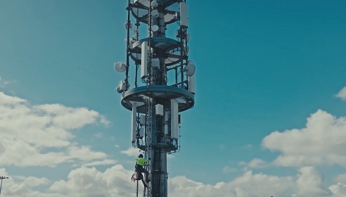 Image of a telecommuncations tower with a man climbing wearing high vis and protective gear, the sky is in the background with clouds.