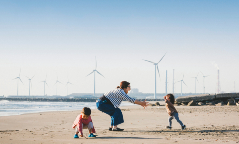 Woman on beach with arms open waiting for her child to run to her with a wind turbine in the background.