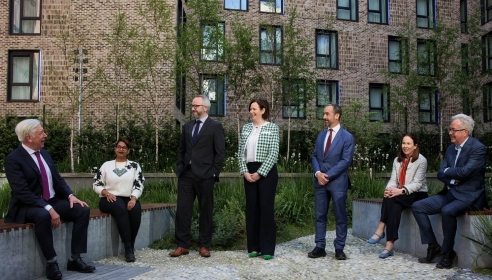 Seven people standing in a courtyard with four sitting and three standing and all looking at the person sitting to the left.