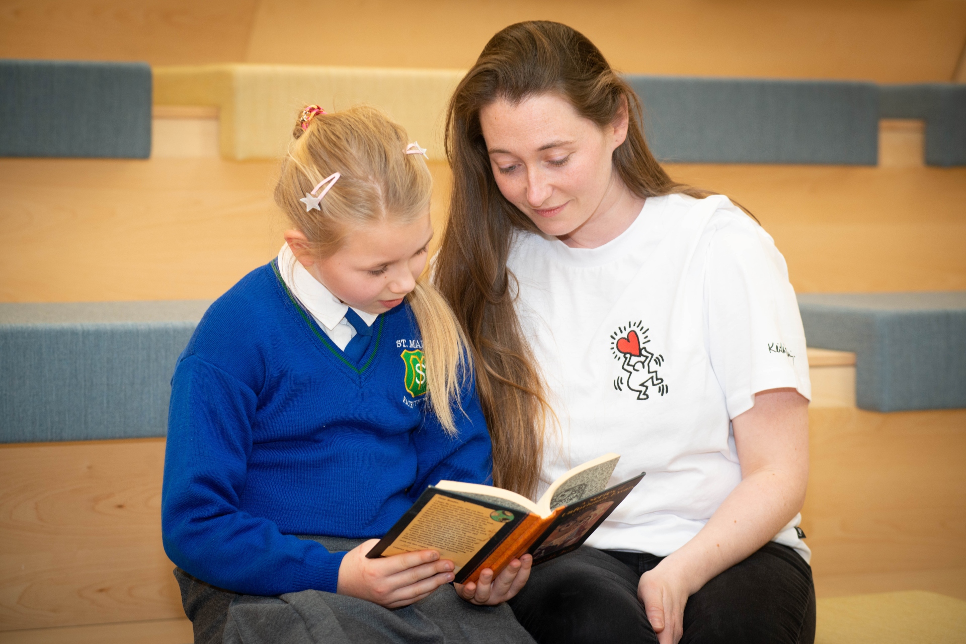 Child with blonde hair and blue school jumper sitting beside a lady with long brown hair and white t-shirt with both looking into a book