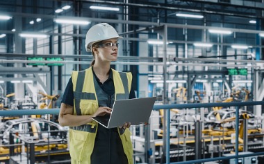 Image of person standing in a factory surveying work with a laptop in their hand.