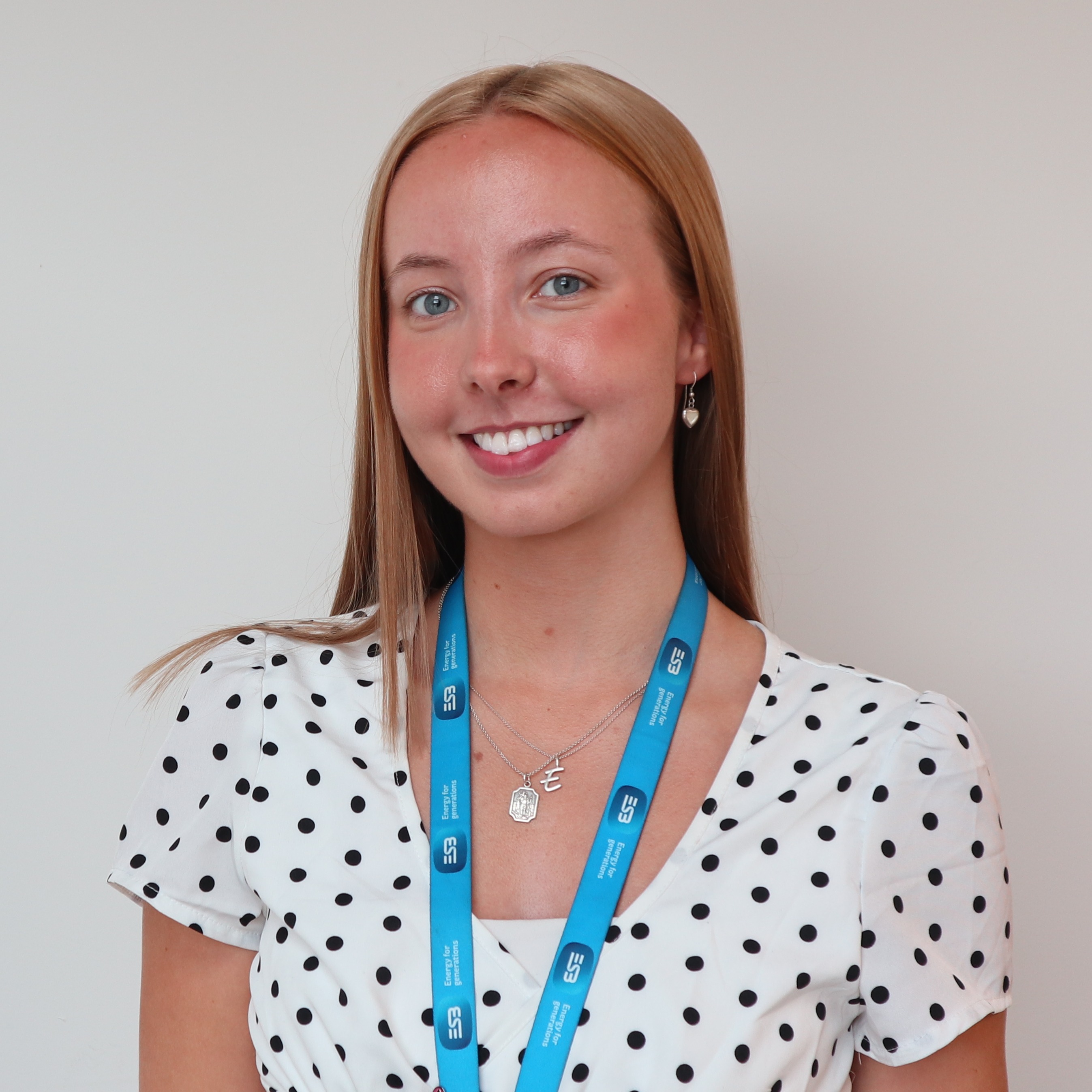 A person stands against a white wall wearing a white top with black polka dots, and a blue lanyard with an ID badge.