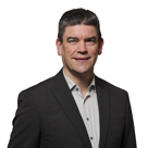 White man with short black hair and wearing a black suit jacket and grey shirt