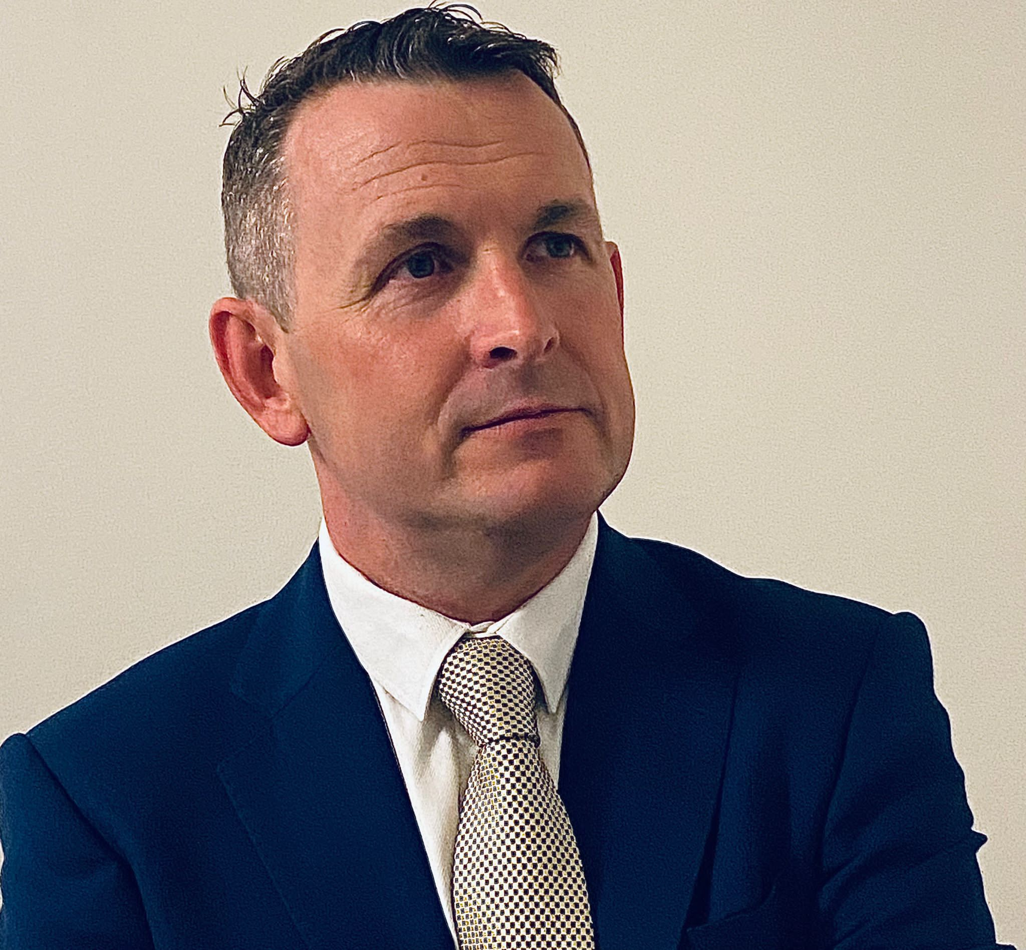 White man with grey hair wearing a blue suit jacket, white shirt and grey tie.