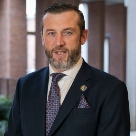 Trevor Walsh, white man with grey/black hair and wearing black suit and white shirt and purple tie