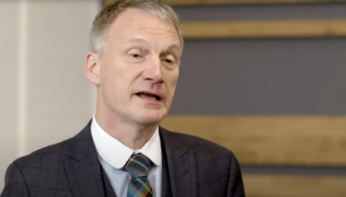 White man with grey hair wearing a black jacket, white shirt and checkered tie.