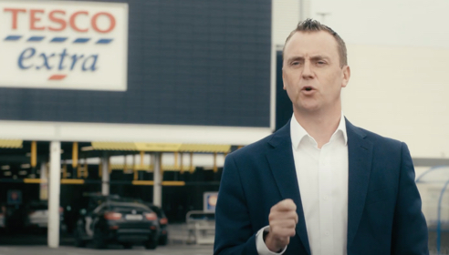 White man wearing a white shirt and blue jacket standing in front of a Tesco Extra shop